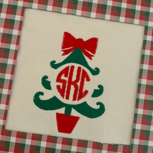 May include: A white fabric with a green and red embroidered Christmas tree design. The tree has a red bow at the top and a red circle with the monogram "SKC" in the center.