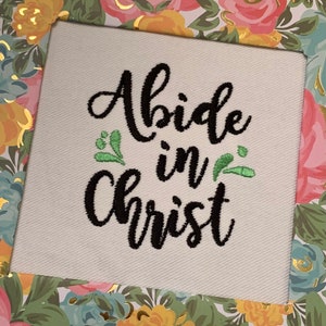 May include: White fabric with black embroidered text that reads "Abide in Christ" with green embroidered leaves on either side of the word "in".