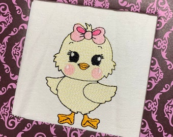May include: Embroidered image of a yellow chick with a pink bow, rosy cheeks, and large black eyes. The chick has orange feet and is standing on a white square. The background is a dark brown with a pink floral pattern.