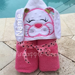 May include: Pink hooded towel with a white bunny face embroidered on the hood. The towel is tied with a black and white polka dot ribbon. The text "Name" is embroidered on the side of the hood.