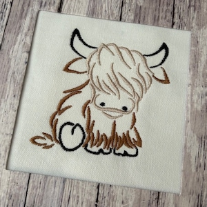 May include: Embroidered design of a brown and white Highland cow with black eyes and hooves. The cow is sitting down with its head down and its tail curled up.
