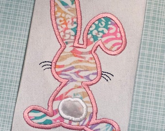 May include: A pink and white embroidered bunny with a white fluffy tail. The bunny is made of fabric with a colorful animal print.