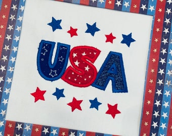 Puede incluir: Una obra de arte enmarcada con la palabra "USA" bordada en rojo, blanco y azul. Las letras están delineadas en azul y rellenas de estrellas. El marco tiene un diseño patriótico con rayas y estrellas rojas, blancas y azules.