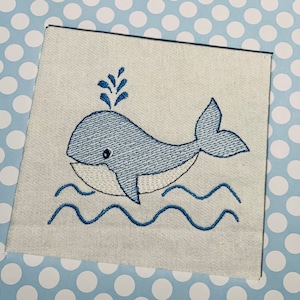 Puede incluir: Diseño bordado de una ballena azul con vientre blanco, nadando en olas azules. La ballena tiene un ojo pequeño y un chorro de agua por encima.