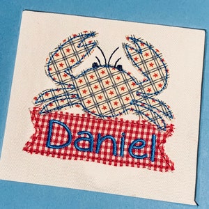 May include: A red and white gingham banner with the name "Daniel" embroidered in blue. A blue and white crab with red stars is above the banner.