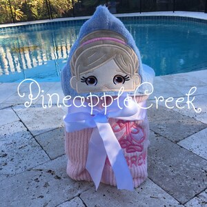 May include: A pink hooded towel with a blue hood featuring an embroidered girl's face. The towel has a white ribbon tied around it and the words "Pineapple Creek" are embroidered on the towel.