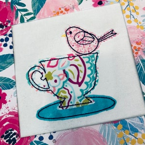 Puede incluir: Una taza de té blanca con un estampado floral colorido y un pájaro rosa posado en el borde. La taza de té está sobre un platillo turquesa.