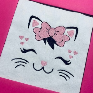 May include: Embroidered design of a white cat face with pink bow, eyelashes, and whiskers. The cat has a pink nose and is surrounded by small pink hearts.