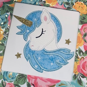 Puede incluir: Un unicornio azul y blanco con los ojos cerrados y un cuerno dorado. El unicornio está bordado sobre un fondo blanco con estrellas doradas.