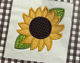 Op de afbeelding: Een geborduurde zonnebloem met gele bloemblaadjes en een bruin centrum met witte stippen. De zonnebloem heeft groene bladeren.