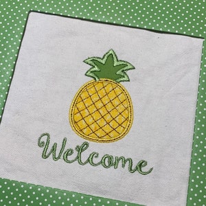 Puede incluir: Una tela blanca con una piña bordada en verde y la palabra "Bienvenido" en bordado verde.
