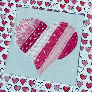 May include: A pink and white fabric heart made of strips of fabric sewn together. The heart is framed in a white frame with a pink and white heart pattern.