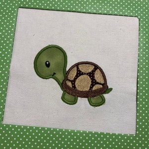 May include: A green and brown embroidered turtle applique. The turtle has a brown shell with a pattern of white circles and a green head and legs.