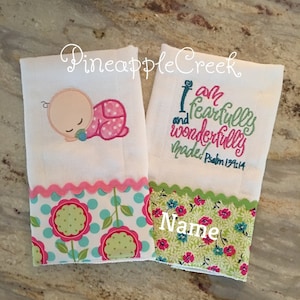 May include: Two white burp cloths with embroidered designs. One cloth features a sleeping baby with a pink and white polka dot onesie and the text "Pineapple Creek". The other cloth features the text "I am fearfully and wonderfully made" with the verse "Psalm 139:14". Both cloths have a pink and green floral print border with the word "Name" embroidered on the bottom.