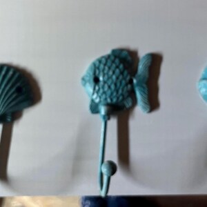 May include: Three blue metal hooks shaped like a seashell, a fish, and a seashell. The hooks are mounted on a white wall.