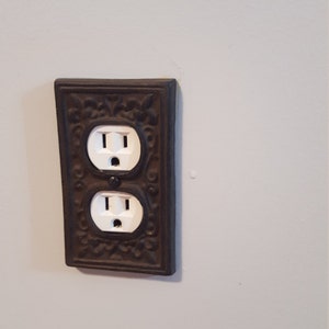 May include: A decorative electrical outlet cover with a dark brown, ornate design. The cover features two standard white electrical outlets. The design includes floral and scrollwork patterns, adding a vintage or antique aesthetic to the wall.
