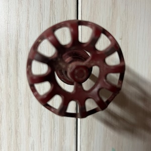 May include: A red metal cabinet knob with a circular design and a wheel-like pattern. The knob has a raised center with a small hole in the middle.