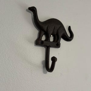 May include: A dark brown cast iron wall hook shaped like a long-necked dinosaur. The dinosaur figure stands on a horizontal bar with two screw holes for mounting, and a single curved hook extends downward from the center of the base.