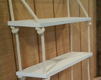 May include: White wooden shelves with three tiers, suspended from the wall with white rope. The shelves are mounted on a wood panel wall.