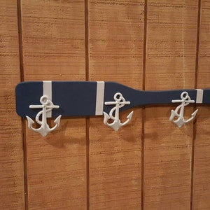 May include: A blue and white painted wooden oar with three white metal anchor hooks attached. The oar is mounted on a wood panel wall.