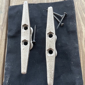 May include: Two silver metal cleat hooks with three holes each. The hooks are on a black fabric background with screws.