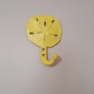 May include: A yellow sand dollar shaped wall hook. The hook is made of metal and has a curved hook at the bottom. The sand dollar has several small holes and a textured surface. The hook is mounted on a light-colored wall.