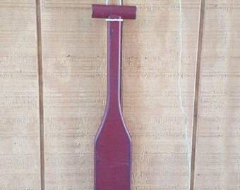 May include: A red wooden paddle with a metal hook at the top, hanging on a wooden wall.