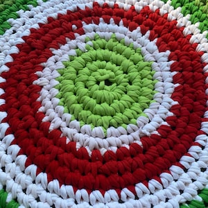 May include: A handmade, circular rug with a spiral design. The rug features concentric rings of green, white, and red fabric. The rug is made of thick, braided material, creating a textured appearance. The rug is suitable for a living room or bedroom.