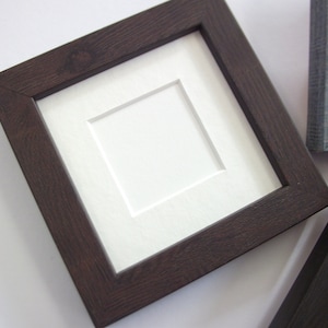 May include: A square, dark brown wooden picture frame with a white mount. The frame has a textured wood grain appearance. Other frames are visible in the background.