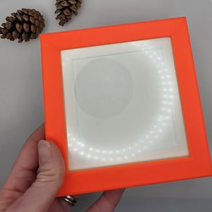 May include: Square orange frame with a white interior and a ring of small lights. The frame is held by a hand. Two brown pinecones are in the background.