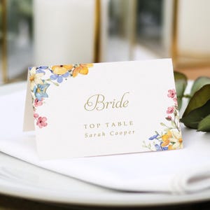 Può includere: Un segnaposto piegato con la parola "Bride" in corsivo dorato. Il biglietto è decorato con composizioni floreali ad acquerello nei toni del giallo, blu e rosa. Il testo "TOP TABLE" e un nome sono stampati sotto.