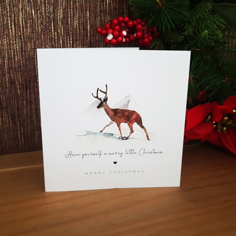May include: A Christmas card featuring a watercolor illustration of a deer walking through a snowy landscape. The card has the text "Have yourself a merry little Christmas. Merry Christmas" printed on it.