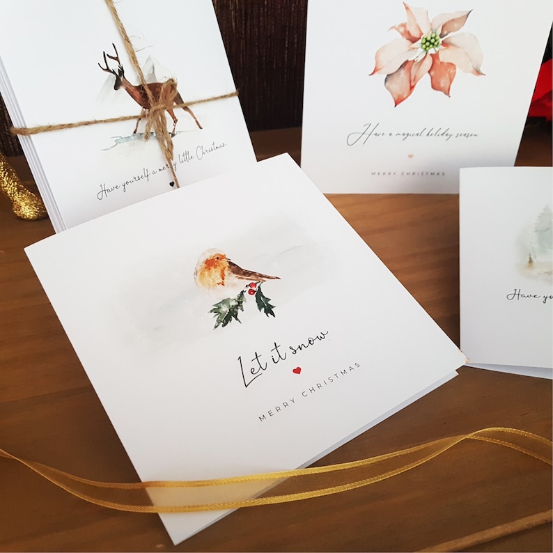May include: Set of four Christmas cards featuring watercolor illustrations. The cards feature a red poinsettia, a deer in a snowy landscape, a robin on a holly branch, and a snowy forest scene. The cards have the text "Have yourself a merry little Christmas", "Have a magical holiday season", "Let it snow", and "Have you..."