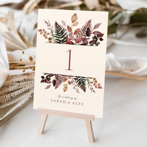 May include: A cream-coloured table number card featuring the number 1 in burgundy. The card is adorned with watercolour botanical illustrations in burgundy, brown, and green. The text "the wedding of SARAH & ALEX" is printed at the bottom. It's displayed on a small wooden easel.
