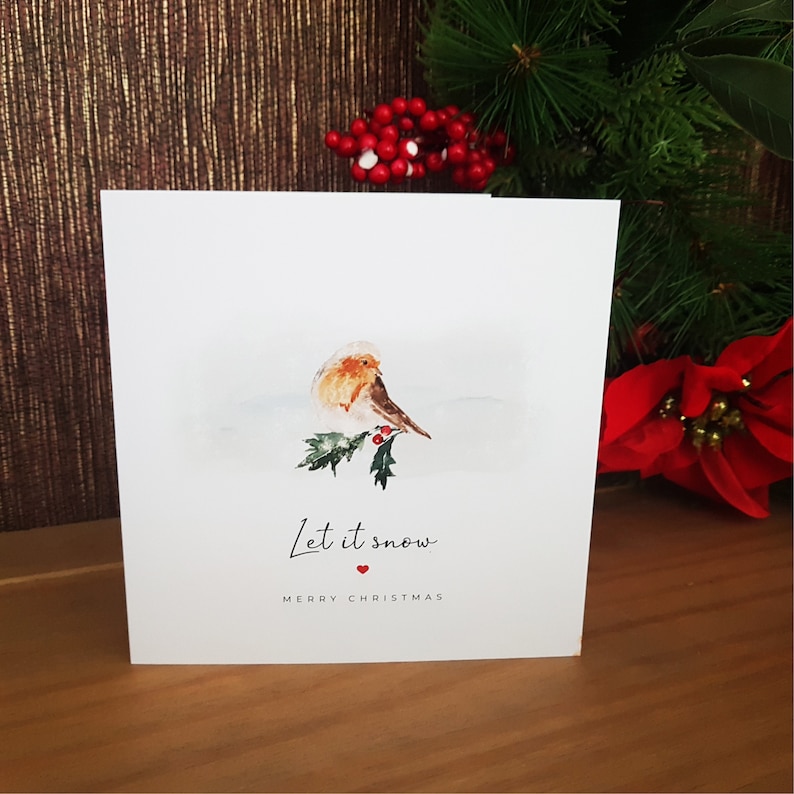 May include: A Christmas card with a watercolor illustration of a robin perched on a holly branch with red berries. The text "Let it snow" is written in cursive with a small red heart below. The card reads "Merry Christmas".
