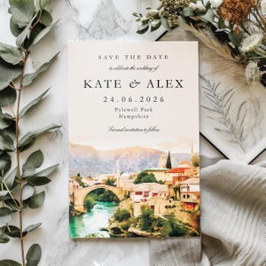 May include: A wedding invitation card with the text "SAVE THE DATE" and the names "KATE & ALEX" along with the date "24.06.2026". The card features a watercolour painting of a bridge over a river and buildings.