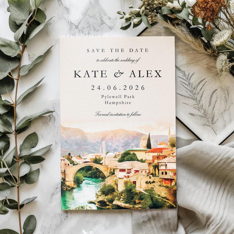 May include: A wedding invitation card with the text "SAVE THE DATE" and the names "KATE & ALEX" along with the date "24.06.2026". The card features a watercolour painting of a bridge over a river and buildings.