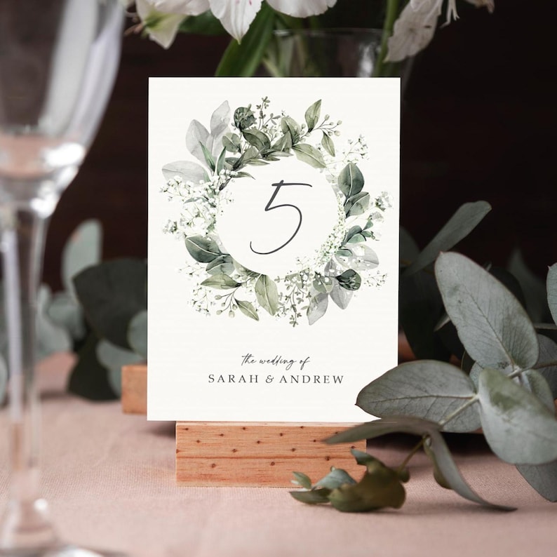 May include: A white card with a green watercolor wreath and the number 5 in the center. The text "the wedding of SARAH & ANDREW" is at the bottom of the card.