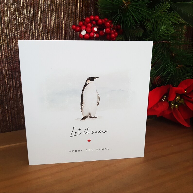 May include: A white Christmas card with a watercolor illustration of a penguin standing on a snowy background. The card says "Let it snow" with a red heart and "Merry Christmas" below.