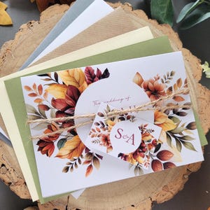 May include: Wedding invitation suite featuring a floral design with autumn colors. The invitation includes the text "The wedding of" and initials "S & A". The suite is tied with twine and includes various colored envelopes.