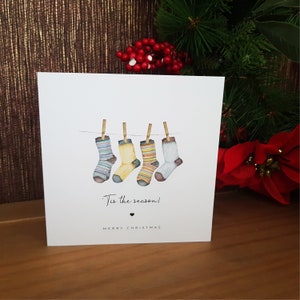 May include: A Christmas card with a watercolor illustration of four colorful socks hanging on a clothesline. The text on the card reads "Tis the season!" and "Merry Christmas".