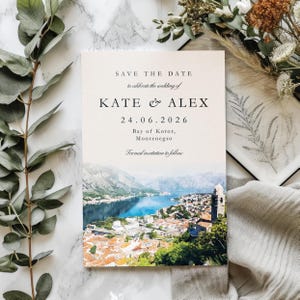 May include: A wedding invitation card with the text "SAVE THE DATE" and the names "KATE & ALEX" along with the date "24.06.2026". The card features a watercolour image of a coastal town and mountains.