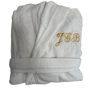 May include: White terry cloth dressing gown with a shawl collar and a matching belt. The gown has gold embroidered initials "JGB" on the chest. The plush fabric is folded neatly, showcasing the design and texture of the garment.