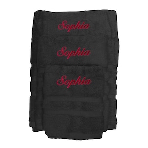 Op de afbeelding: Een stapel van drie zwarte badhanddoeken met de naam "Sophia" in rood script geborduurd. De handdoeken zijn opgevouwen en gestapeld, waarbij de gepersonaliseerde borduursels zichtbaar zijn.