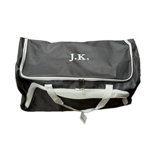 May include: A black duffel bag with a white zipper and a white mesh panel. The bag has a white handle and the initials "J.K." embroidered on the front.