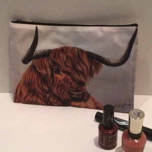 May include: A white zippered makeup bag with a brown and white Highland cow illustration. The bag is on a white surface with two bottles of nail polish and a black eyeliner.