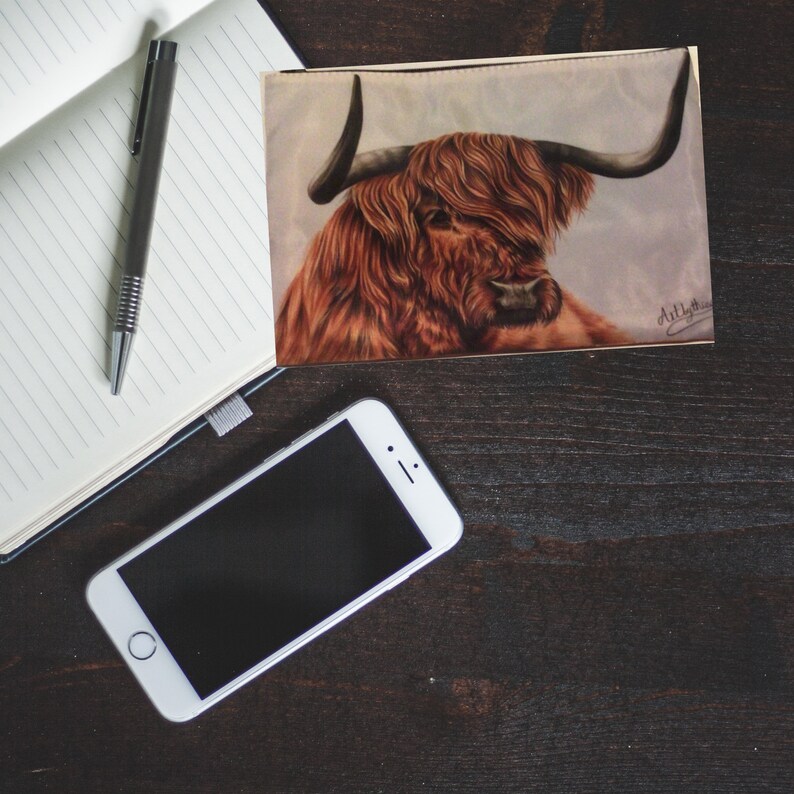 May include: A close-up of a brown and white Highland cow with long horns and a shaggy mane. The cow is painted on a white background with a light blue sky. The painting is signed "Art by Jess".