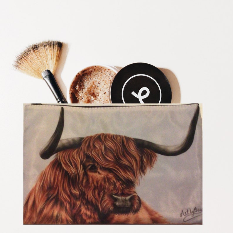 May include: A beige zippered pouch with a brown and white Highland cow illustration. The cow has long, shaggy hair and a black nose. The pouch has a white background with a subtle gray pattern. The text "Artlythis" is written in black in the bottom right corner.
