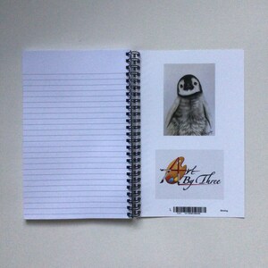 May include: Spiral-bound notebook with lined pages. The right page features a pencil sketch of a penguin and a logo that reads "Art By Three". The notebook is open, showing the spiral binding and the blank pages.