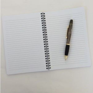 May include: An open, lined notebook with a black spiral binding and a pen. The pen is dark gray with gold accents and a black grip. The notebook's pages are white with horizontal lines.